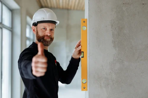 Professional construction worker with hard hat and spirit level giving thumbs up indoors.